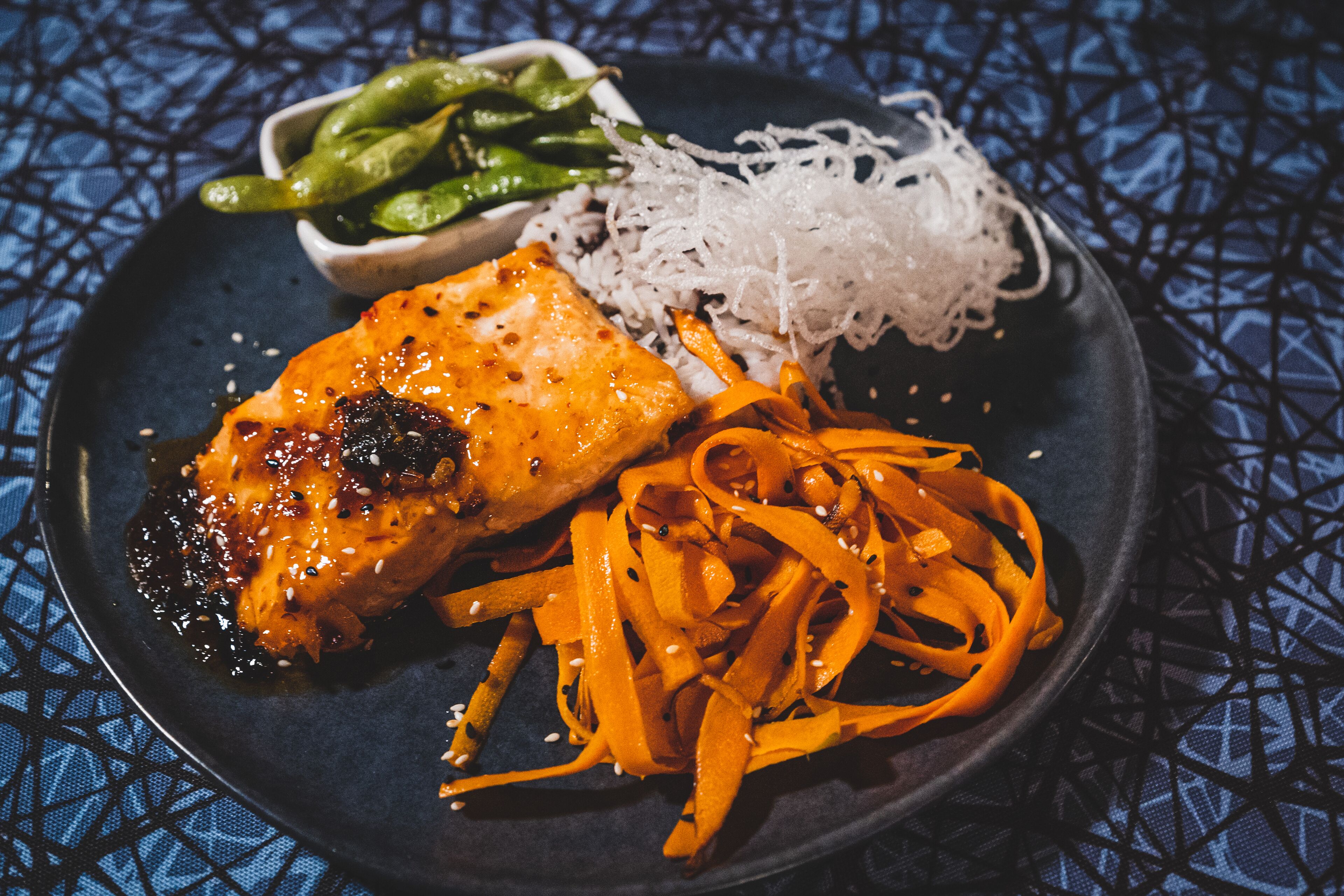 A plate with grilled salmon, shredded carrots, and green beans on a dark plate with a blue patterned tablecloth.