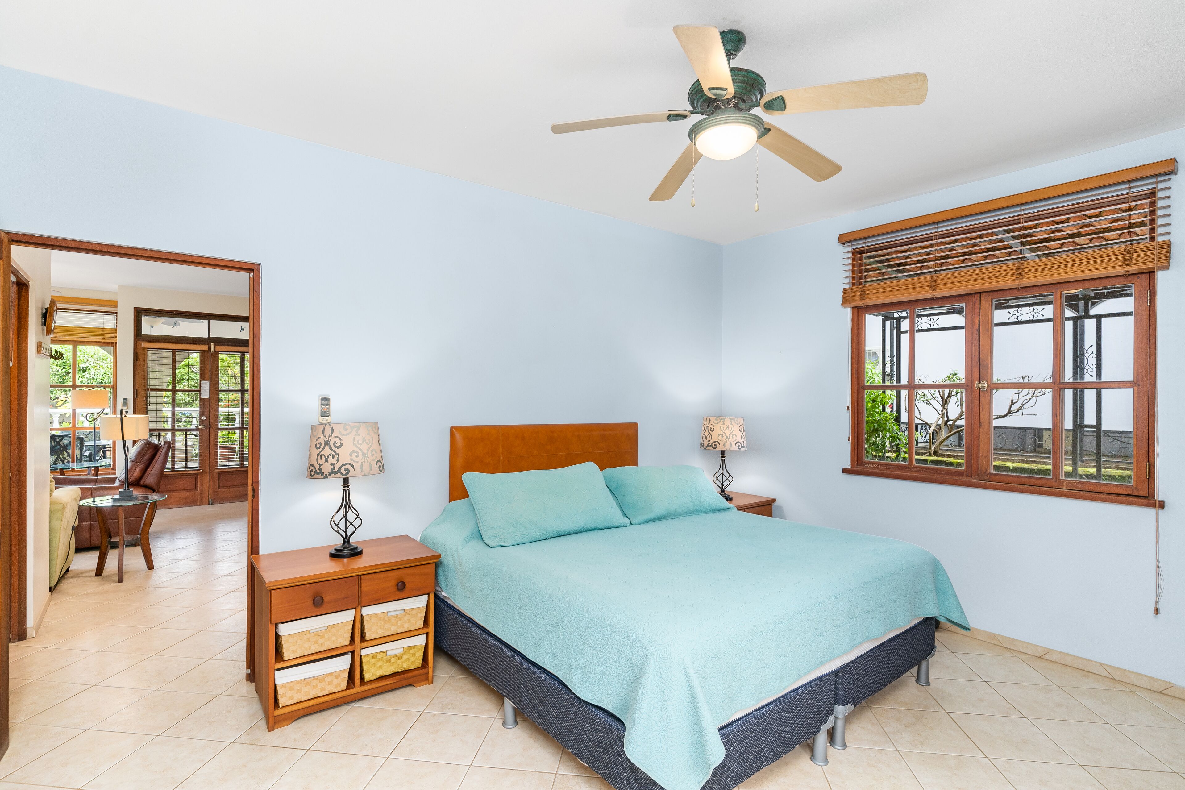 Un dormitorio con cama, mesitas de noche de madera, ventilador de techo y ventana con persianas.