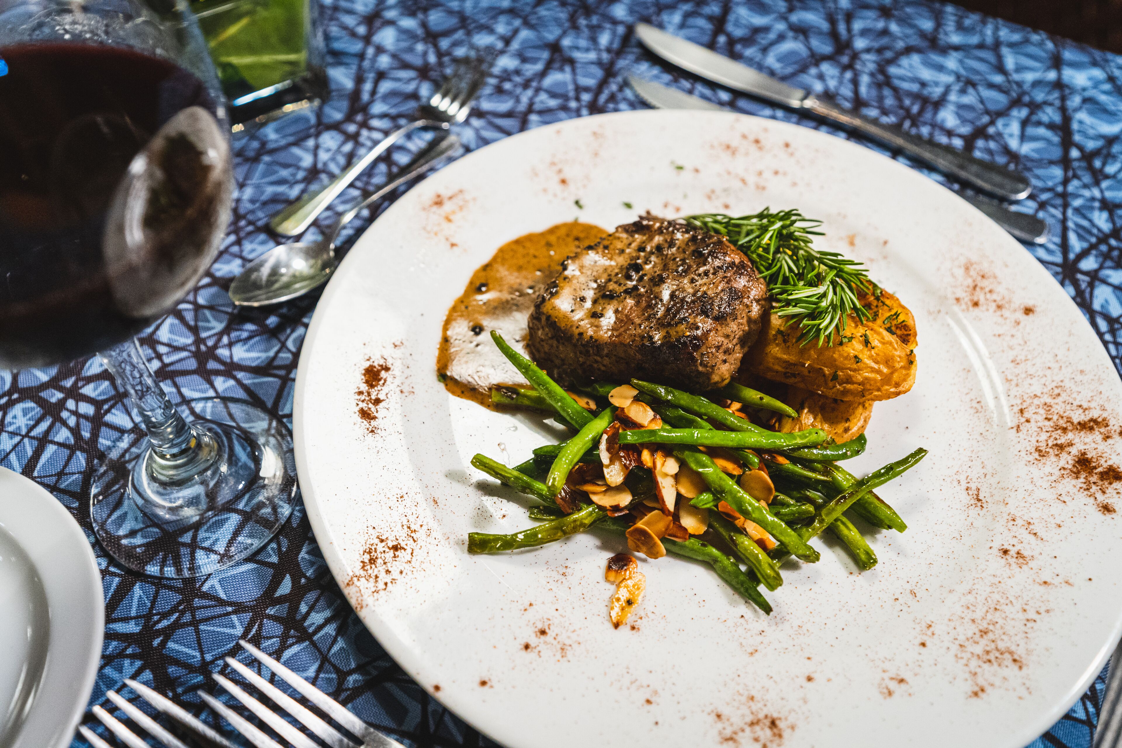 Een bord met een steak, groene bonen en een aardappel, vergezeld van een glas rode wijn op een tafel met een blauw geruit tafelkleed.