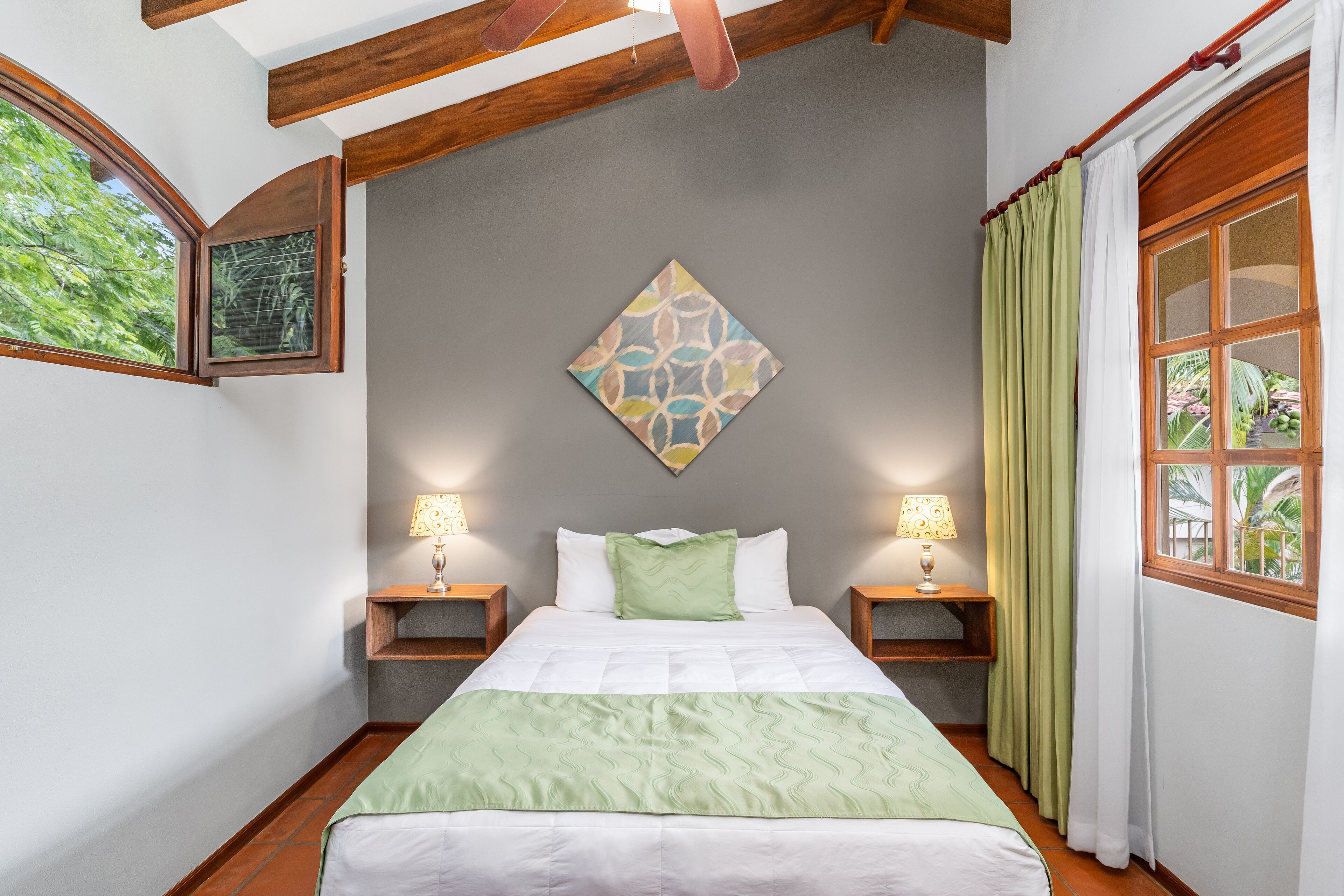 Un dormitorio con cama, dos mesitas de noche, una lámpara de pared, una ventana con cortinas e imagen enmarcada en la pared.