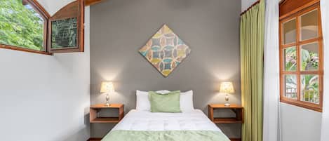 A bedroom with a bed, two bedside tables, a wall-mounted lamp, a window with curtains, and a framed picture on the wall.
