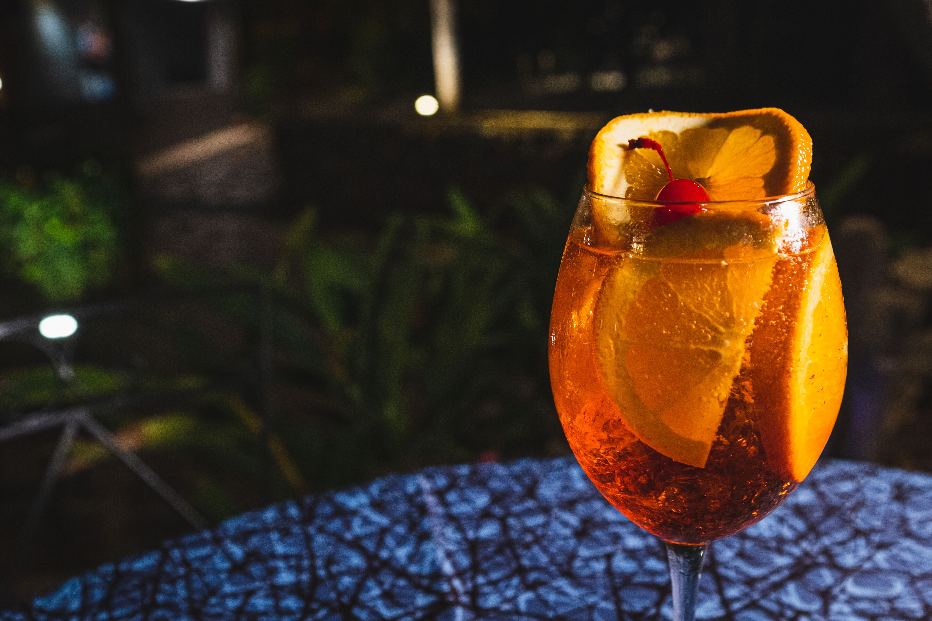 A glass of drink with a slice of orange and a cherry on a table.