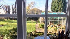 Blick durch ein Fenster auf einen Gartenweg, einen Teich und ein Gewächshaus.