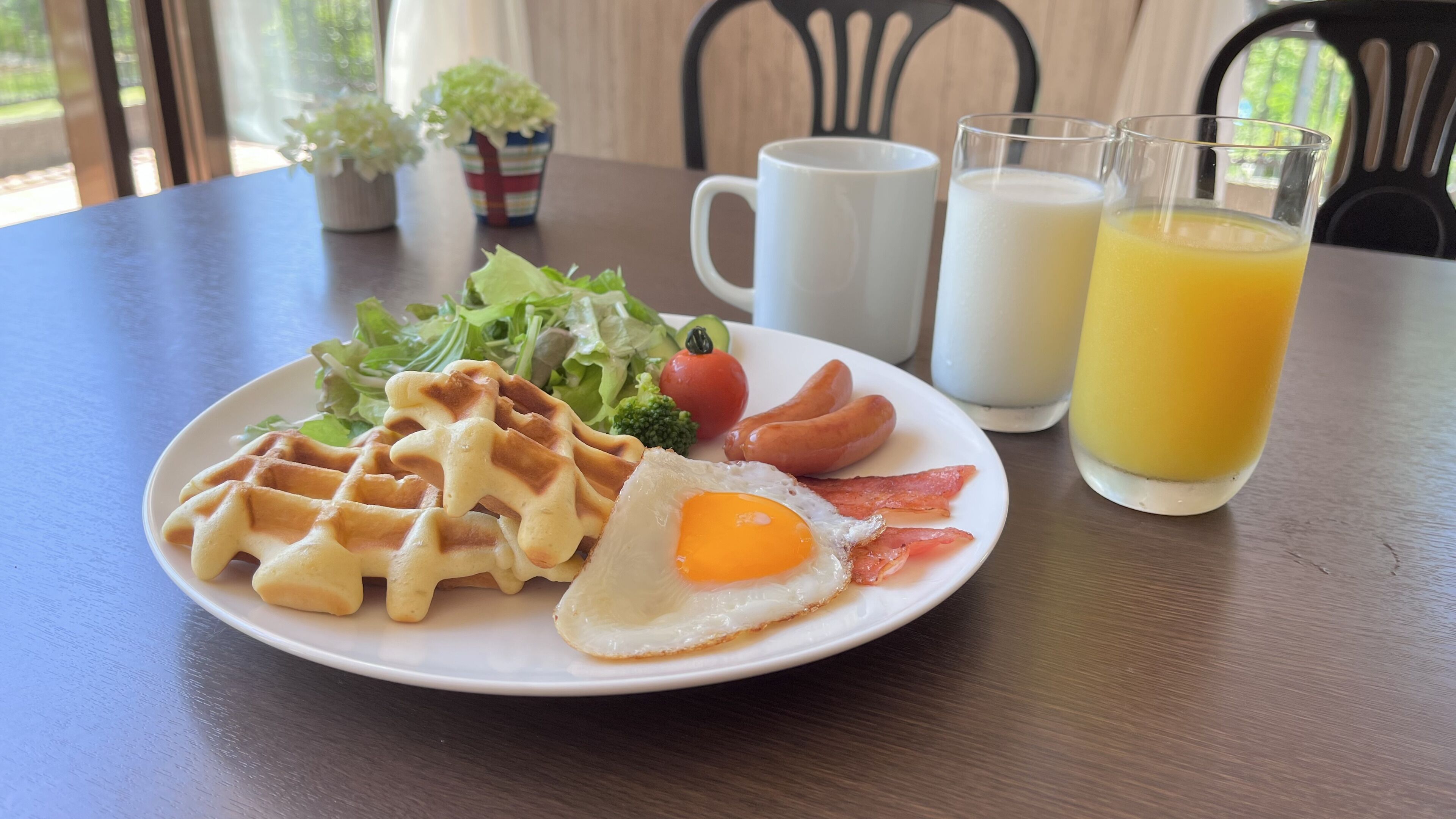 A plate with waffles, a fried egg, bacon, and vegetables, accompanied by a cup of coffee and a glass of orange juice on a wooden table.