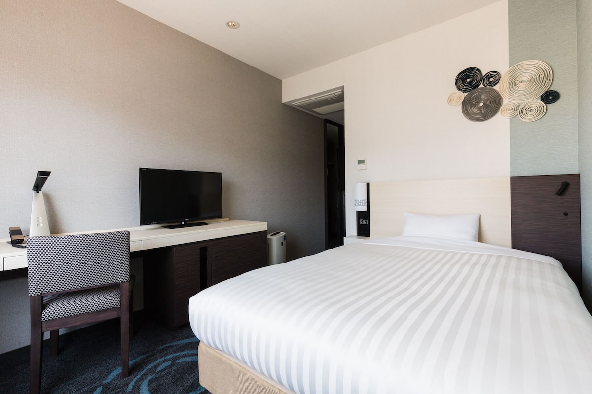 Habitación de hotel con cama, escritorio con televisor, una silla y una obra de arte en la pared.