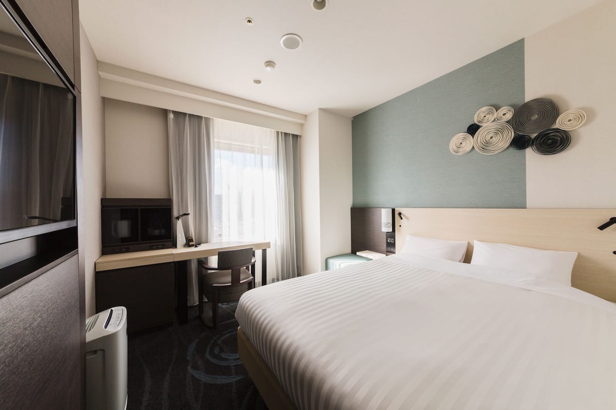 Habitación de hotel con cama, escritorio, silla, televisor y reloj en la pared.