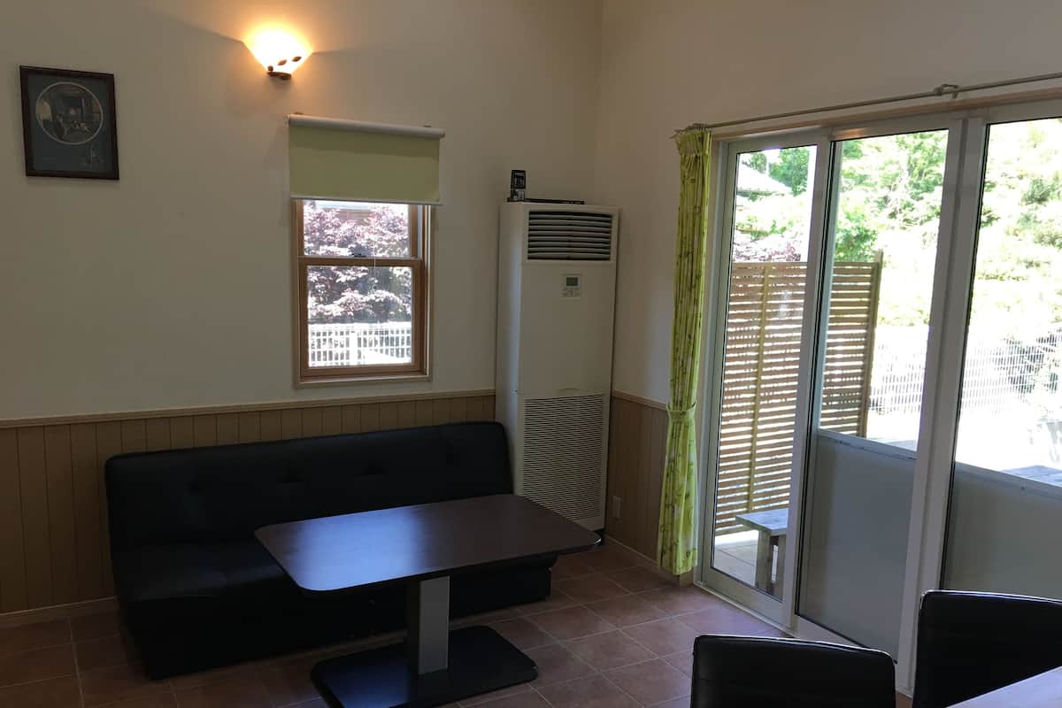 A small room with a black sofa, a table, a window with a view of trees, and an air conditioning unit.