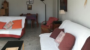 Een woonkamer met een bank, een stoel, een tafel en een lamp. Achterin staat een bed.