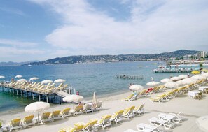 A beach with rows of sun loungers and umbrellas, a pier extending into the water, and a cityscape in the background.