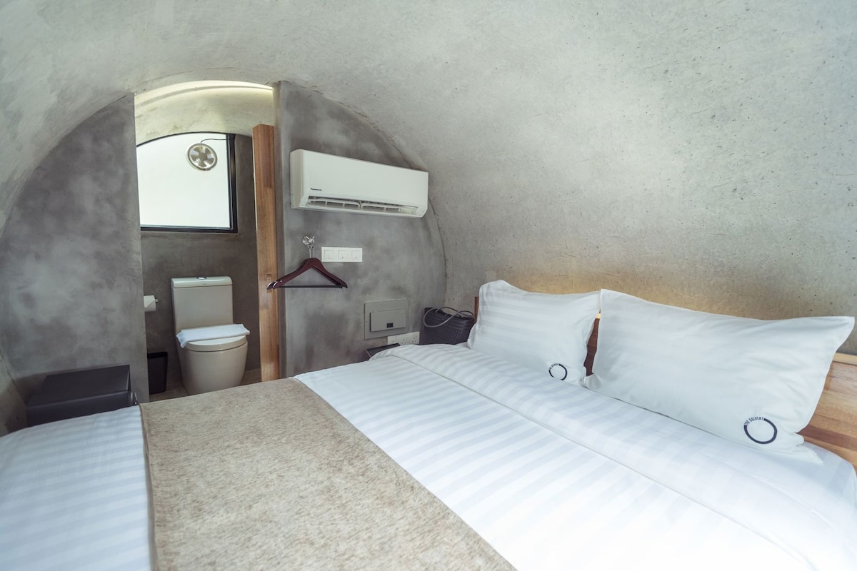 Habitación compacta con cama, inodoro suspendido y equipo de aire acondicionado.