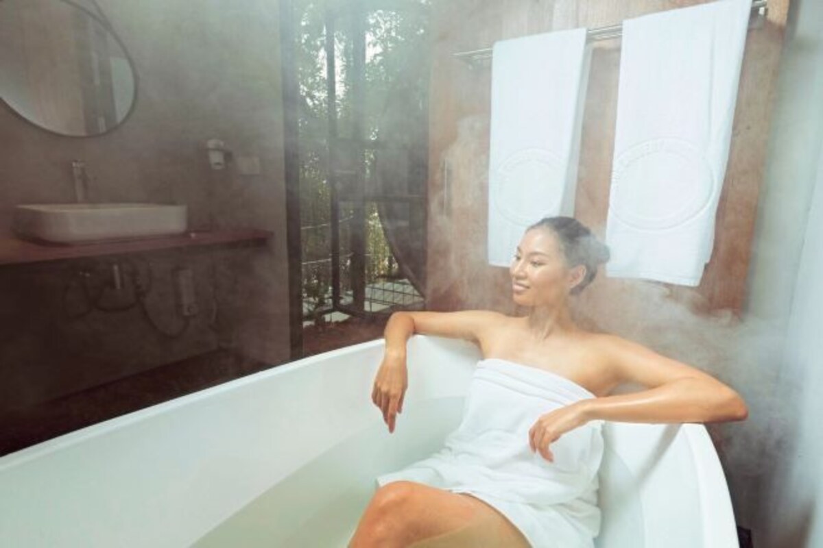 A person in a bathtub with steam, a sink, and a mirror on the wall.