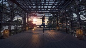 A person performing a yoga pose on a wooden deck with a glass ceiling, overlooking a body of water and trees. - The Culvert (Kuching)