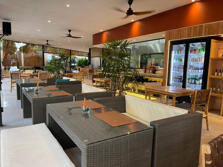 Un área de estar moderna al aire libre con mobiliario de mimbre y mesas de vidrio, una zona de bar con un refrigerador lleno de bebidas y ventiladores de techo.