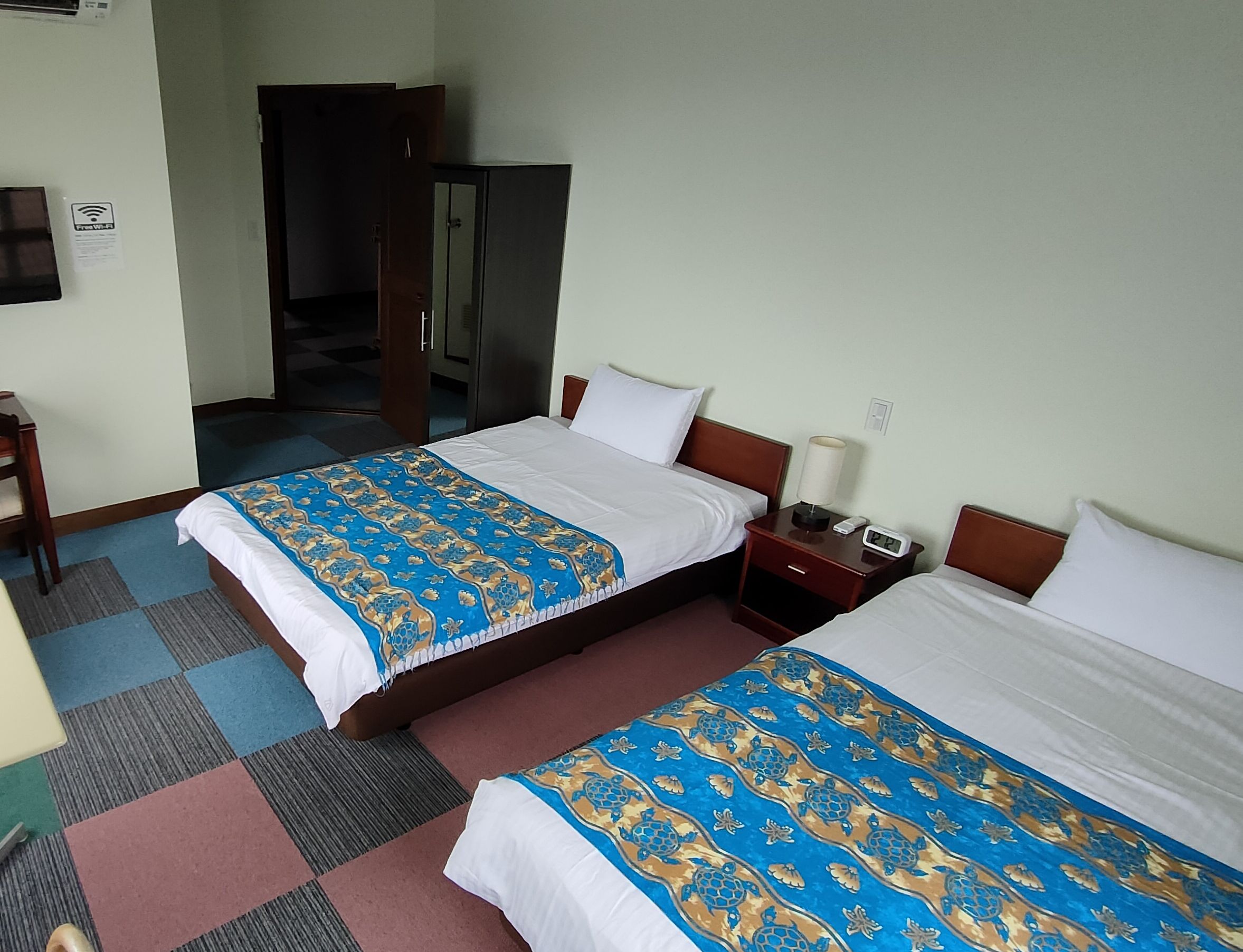Une chambre d’hôtel avec deux lits, une table de chevet avec une lampe et une horloge, un bureau et une chaise.
