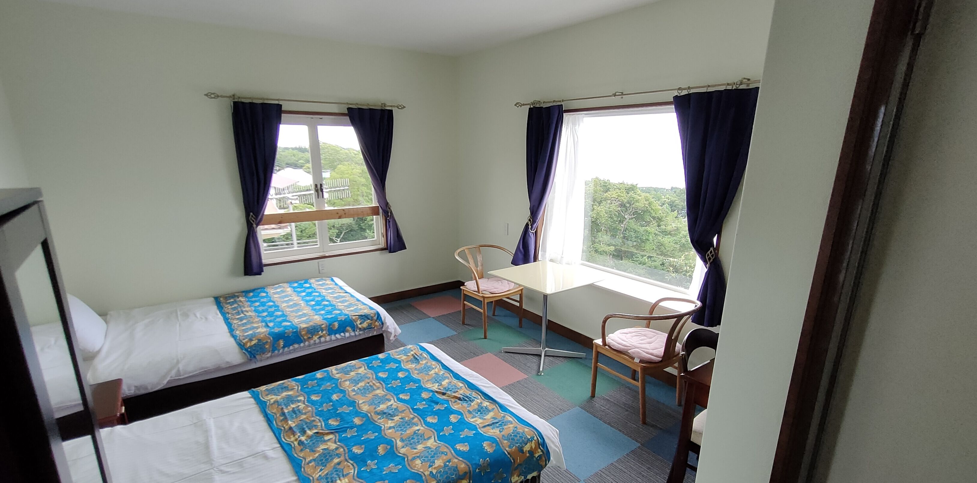 A room with two beds, a table, and chairs, with windows offering a view of greenery.