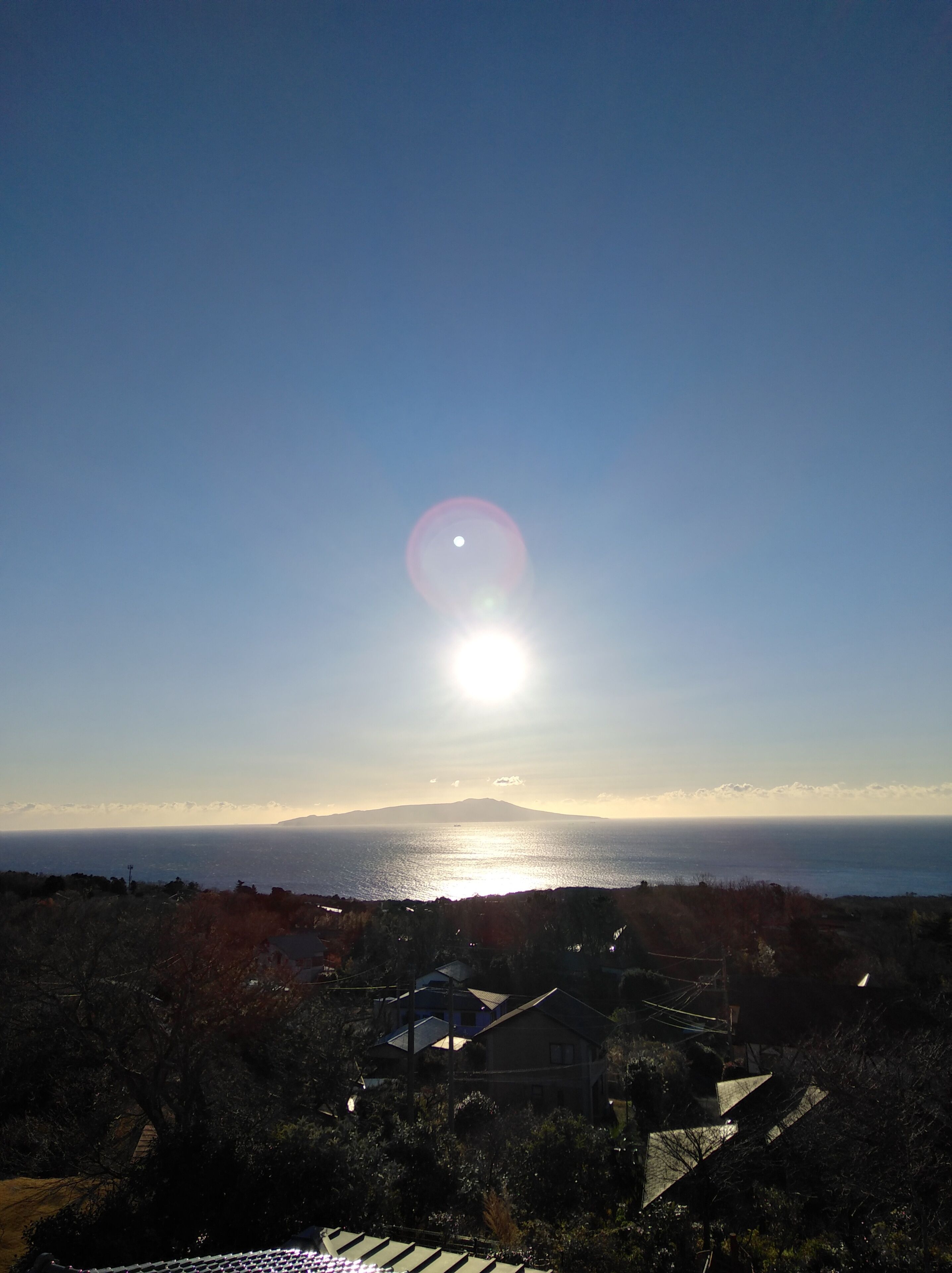 A coastal view with a clear sky, a sun reflection on the water, and a distant mountain.