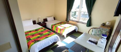 A hotel room with two beds, a small table with a chair, a window with curtains, and a wall-mounted lamp.