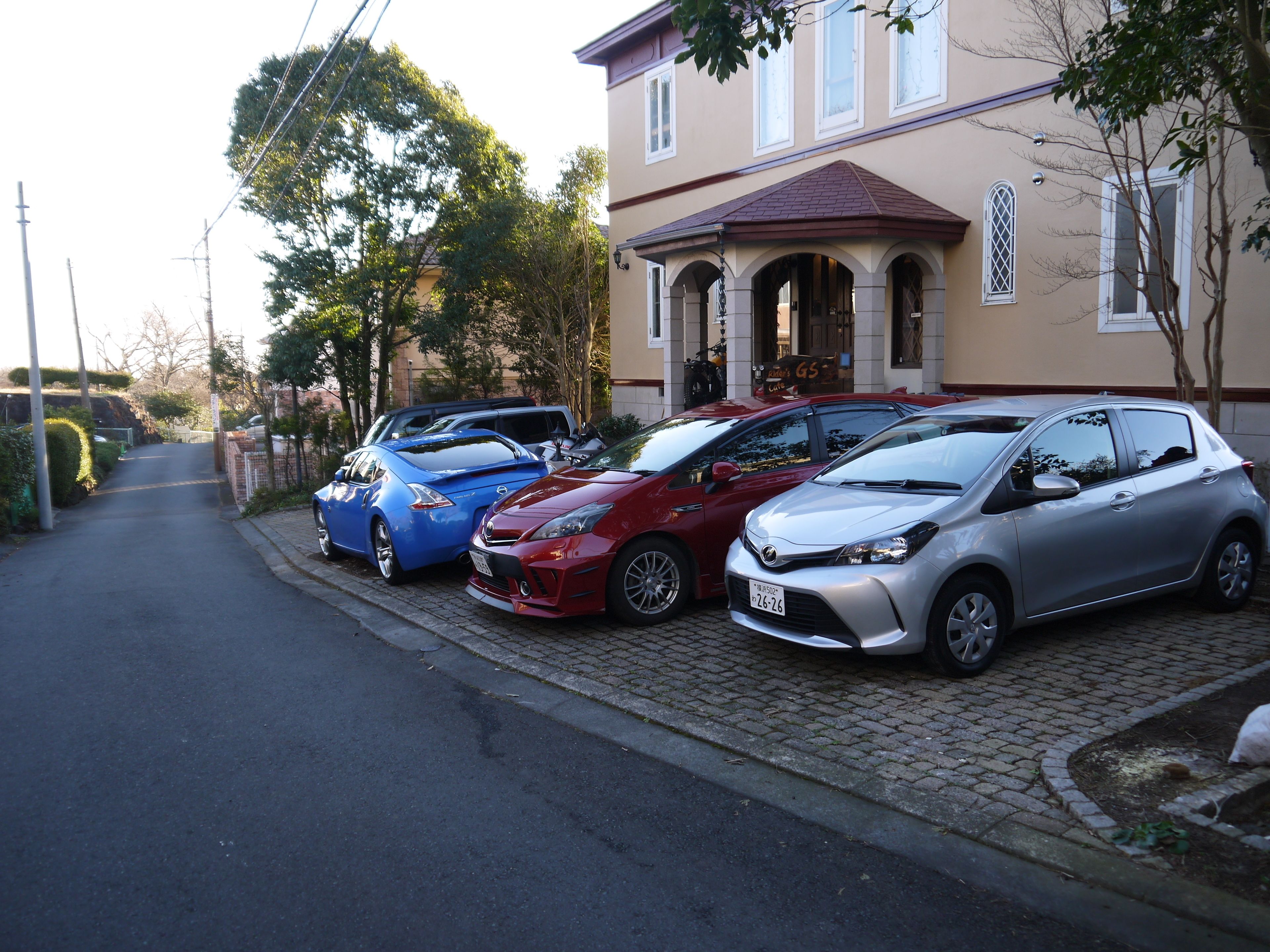 Una hilera de coches estacionados a lo largo de una calle empedrada en una zona residencial, con un edificio que presenta ventanas de arco y un tejado de tejas.