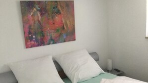 A neatly made bed with white bedding and pillows, a colorful abstract painting on the wall, and a small bedside table with a lamp.