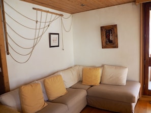 Een hoekwoonkamer met een beige modulaire bank, twee gele kussens, een lijst met een afbeelding, en een houten plafond.
