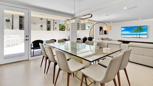 A modern dining area with a glass table surrounded by chairs, a sofa, and a TV displaying a beach scene. - 5BR/5BA Large Family Luxury Beachfront Condo, Private Terrace & Pool (South Padre Island)