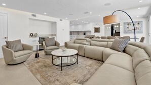 A modern living room with a beige sectional sofa, two armchairs, a coffee table, and a kitchen in the background. - 5BR/5BA Large Family Luxury Beachfront Condo, Private Terrace & Pool (South Padre Island)