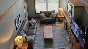 Ein Loft-Wohnzimmer mit hoher Decke, zwei großen Fenstern, einer Sofagarnitur, einem Couchtisch und einem Fernseher.