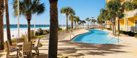 A pool area with a view of the beach and a pier, surrounded by palm trees and benches.