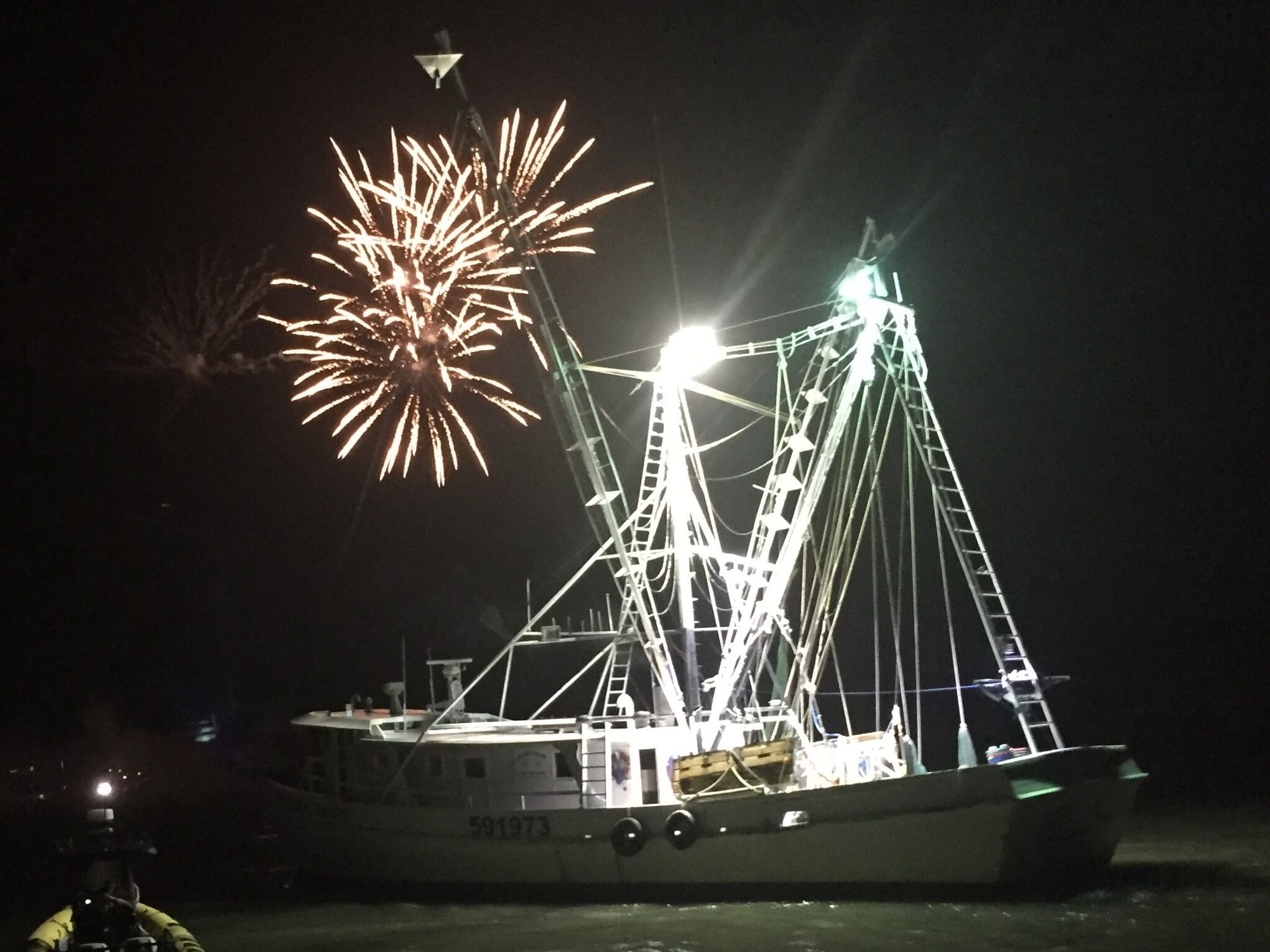 A boat with a crane and a ladder, docked at night with fireworks in the background.