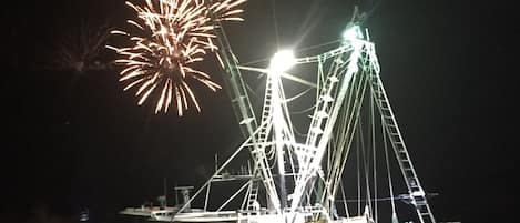A boat with a crane and a ladder, docked at night with fireworks in the background.