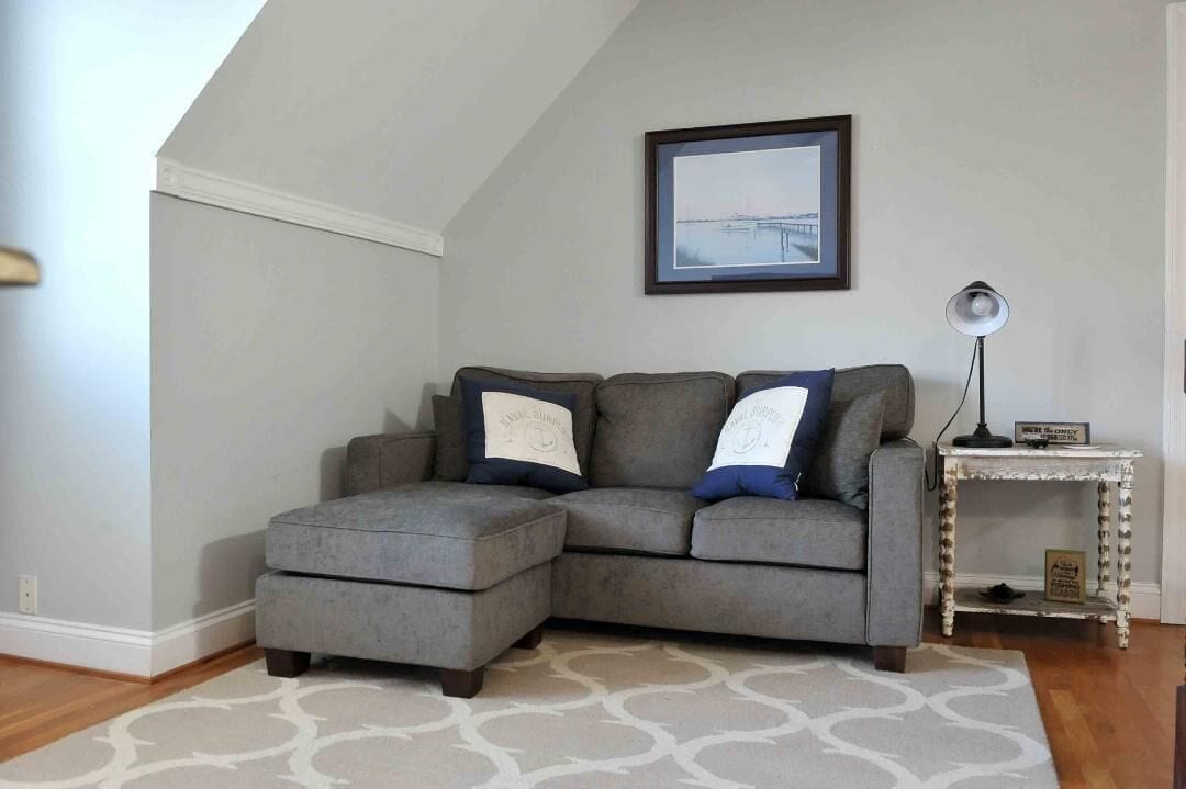A gray sectional sofa with matching throw pillows, a framed picture on the wall, a small white side table, and a patterned rug.