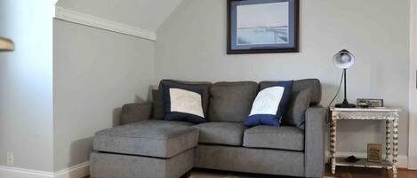 A gray sectional sofa with matching throw pillows, a framed picture on the wall, a small white side table, and a patterned rug.