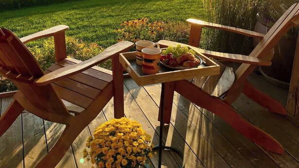 A wooden deck with two chairs and a table set for a meal, overlooking a grassy field and mountains.