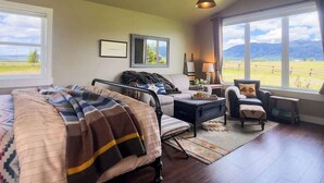 A cozy living room with a bed, sofa, and armchair, large windows with a view of the outdoors, and a ceiling fan.