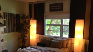 Camera da letto con un letto, lampade sui comodini, una finestra vista alberi, una pianta e uno scaffale con libri.