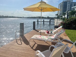 A wooden deck with lounge chairs and a yellow umbrella, overlooking a body of water with a dock and buildings in the background. - Premium Beachside Get-Away (Ft Lauderdale)
