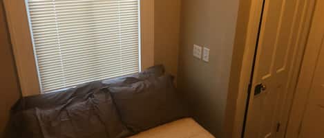Une petite chambre avec un lit, une fenêtre avec des stores, une porte et un interrupteur mural.