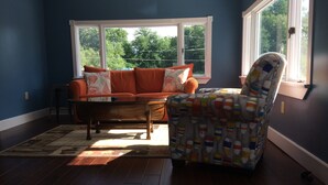 A living room with a blue wall, a wooden floor, a sofa with orange cushions, a patterned armchair, and a window with a view of trees. - Amazing view of Seneca Lake, close to wine trail and water activities (Geneva)