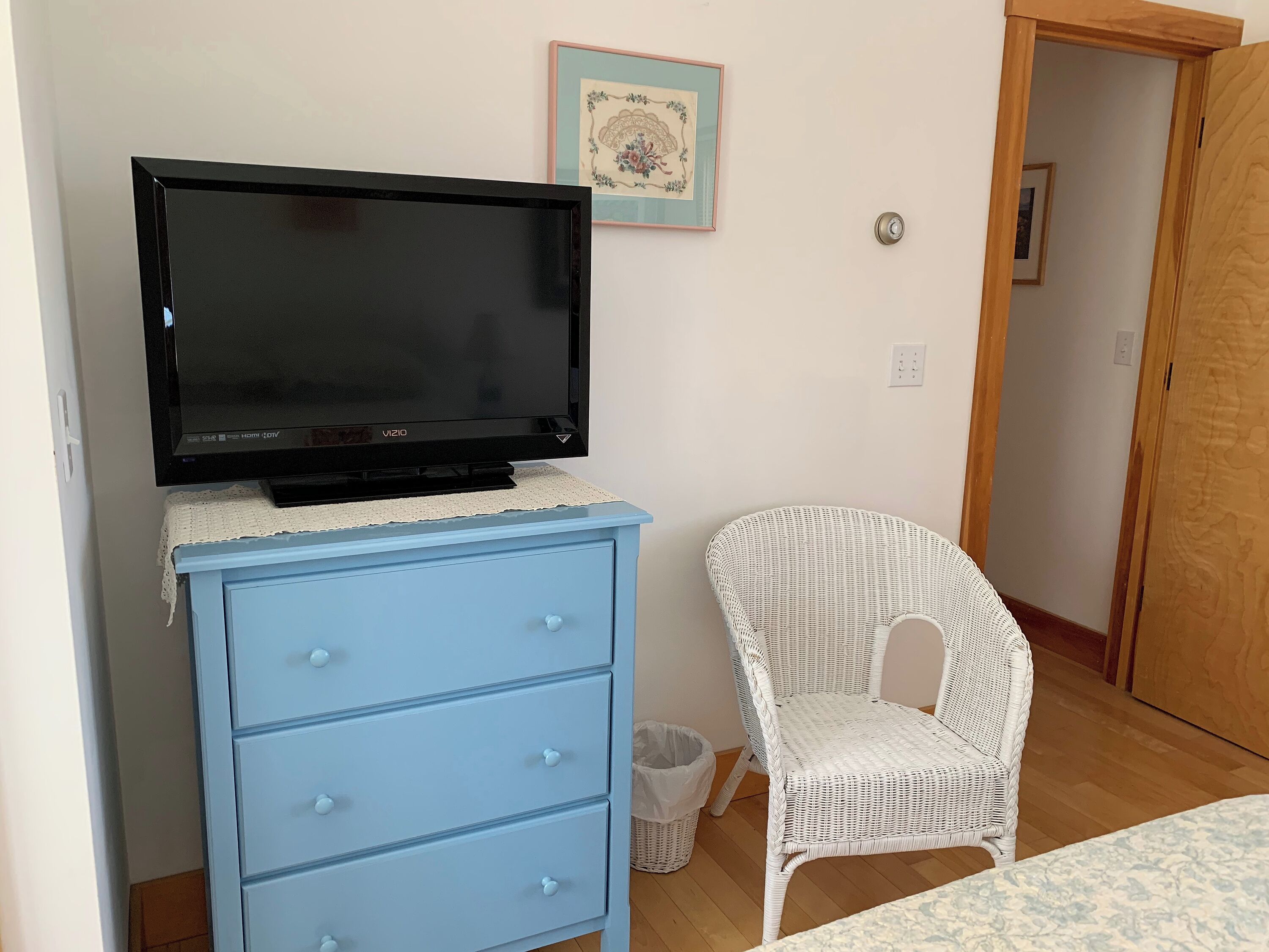 A room with a TV on a blue dresser, a wicker chair, a small trash bin, and a door leading to another room.