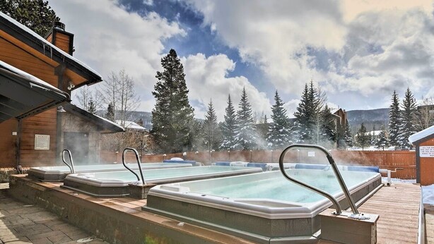 Un espace dédié aux bains à remous, comprenant trois bains à remous, entouré d’une terrasse en bois et d’arbres couverts de neige.