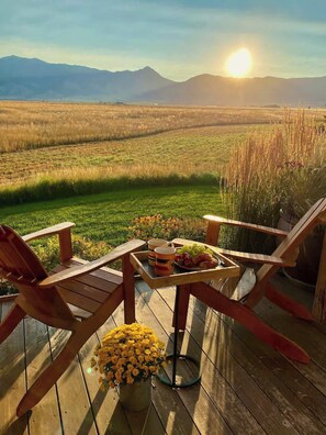 Una terraza de madera con dos sillas y una mesa lista para una comida, con vista a un campo cubierto de hierba y montañas.