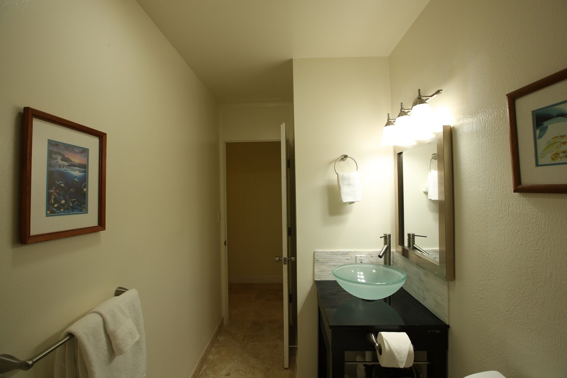 A bathroom with a glass sink, a towel rack, a framed picture on the wall, and a door leading to another room.