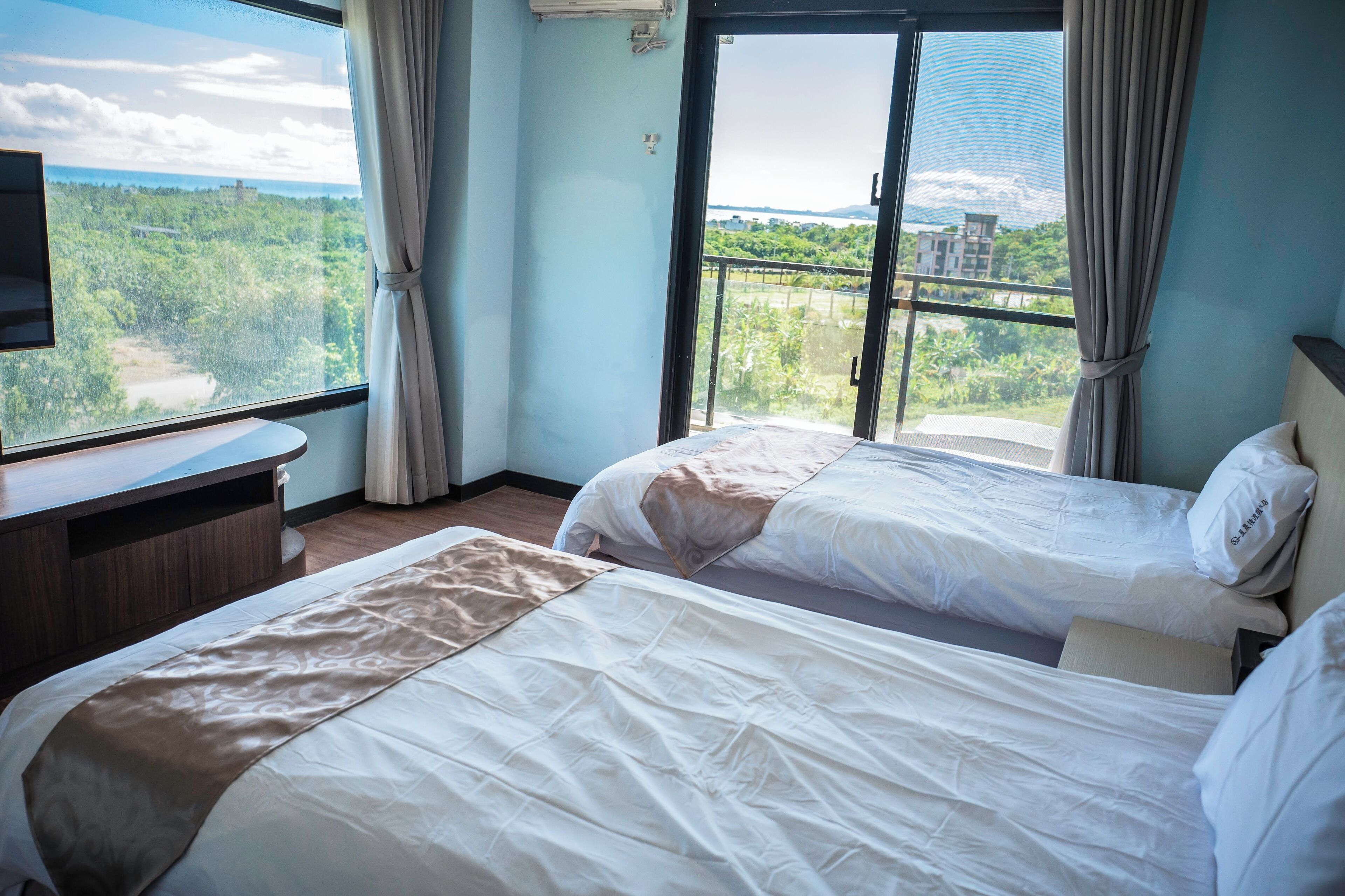 Habitación de hotel con dos camas, televisión y balcón con vistas a zonas verdes y edificios.