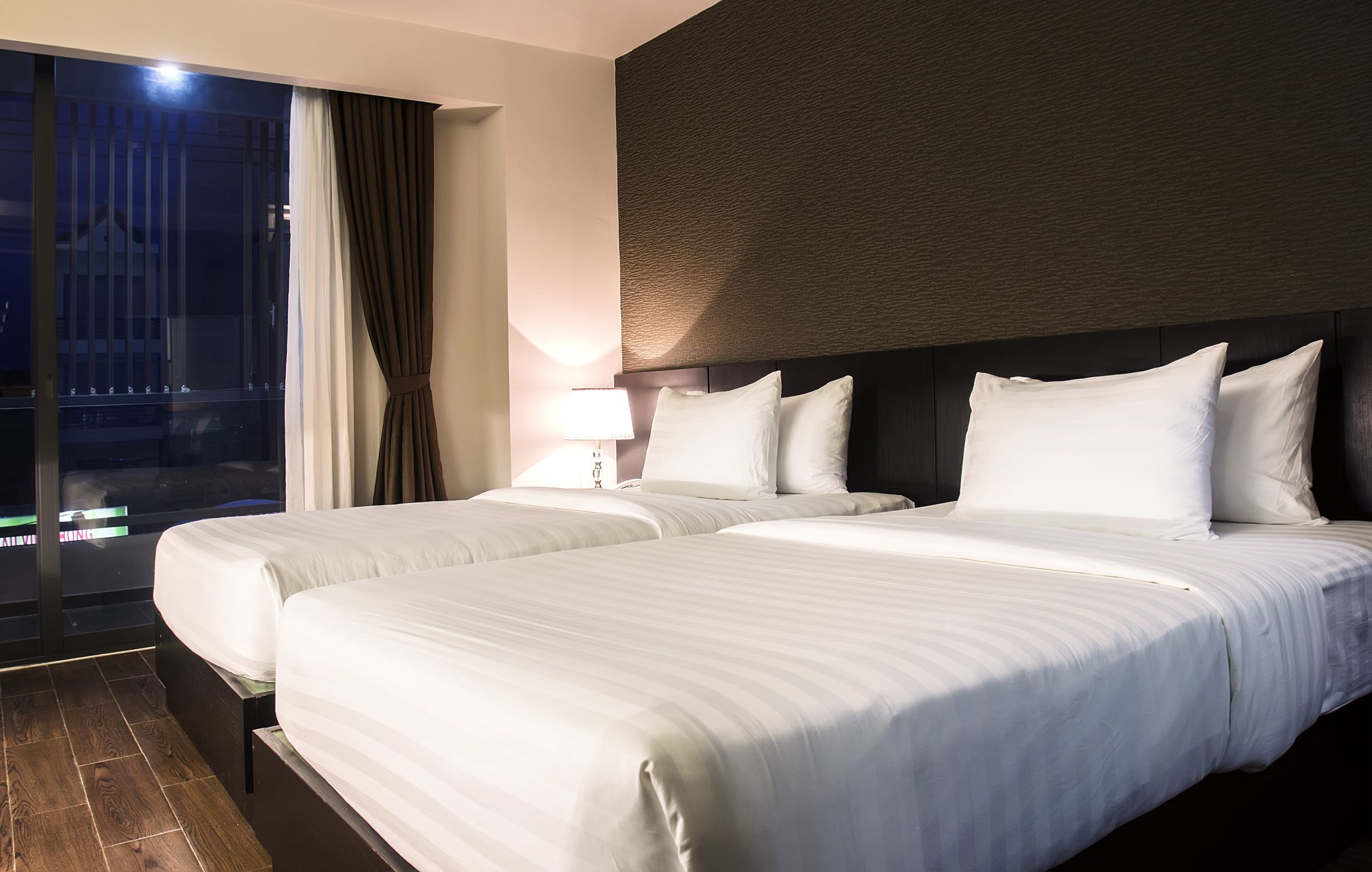 A hotel room with two beds, a large window with curtains, a wall-mounted headboard, and a bedside lamp.