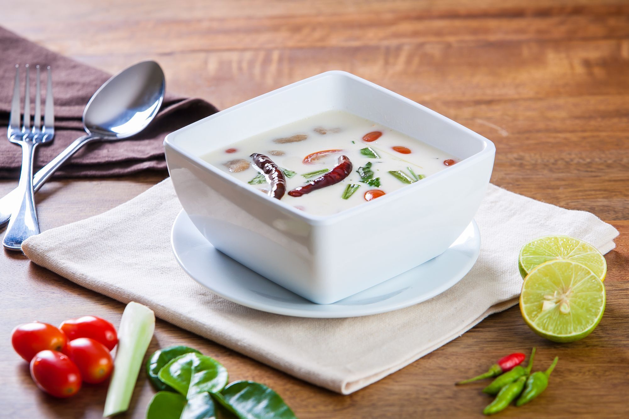 A bowl of soup on a white plate with a spoon and fork beside it, placed on a wooden table with a cloth napkin.