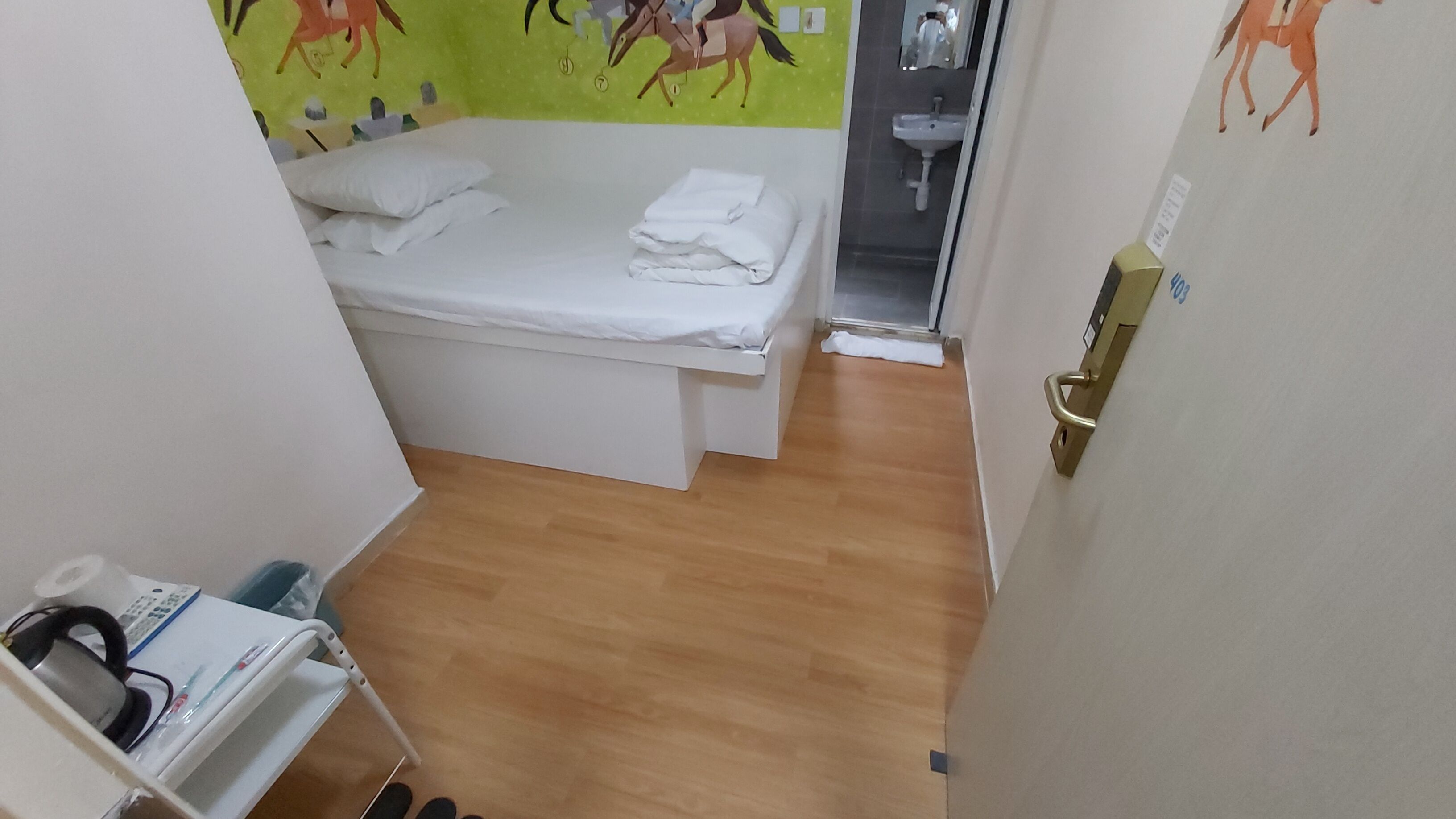 A small, single-bed room with a wooden floor, a white bed with pillows, a small table with a chair, and a wall with a horse-themed mural.