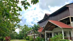 A large building with a red roof and multiple balconies, surrounded by lush greenery and palm trees. - RTMS Guesthouse Semporna (Semporna)