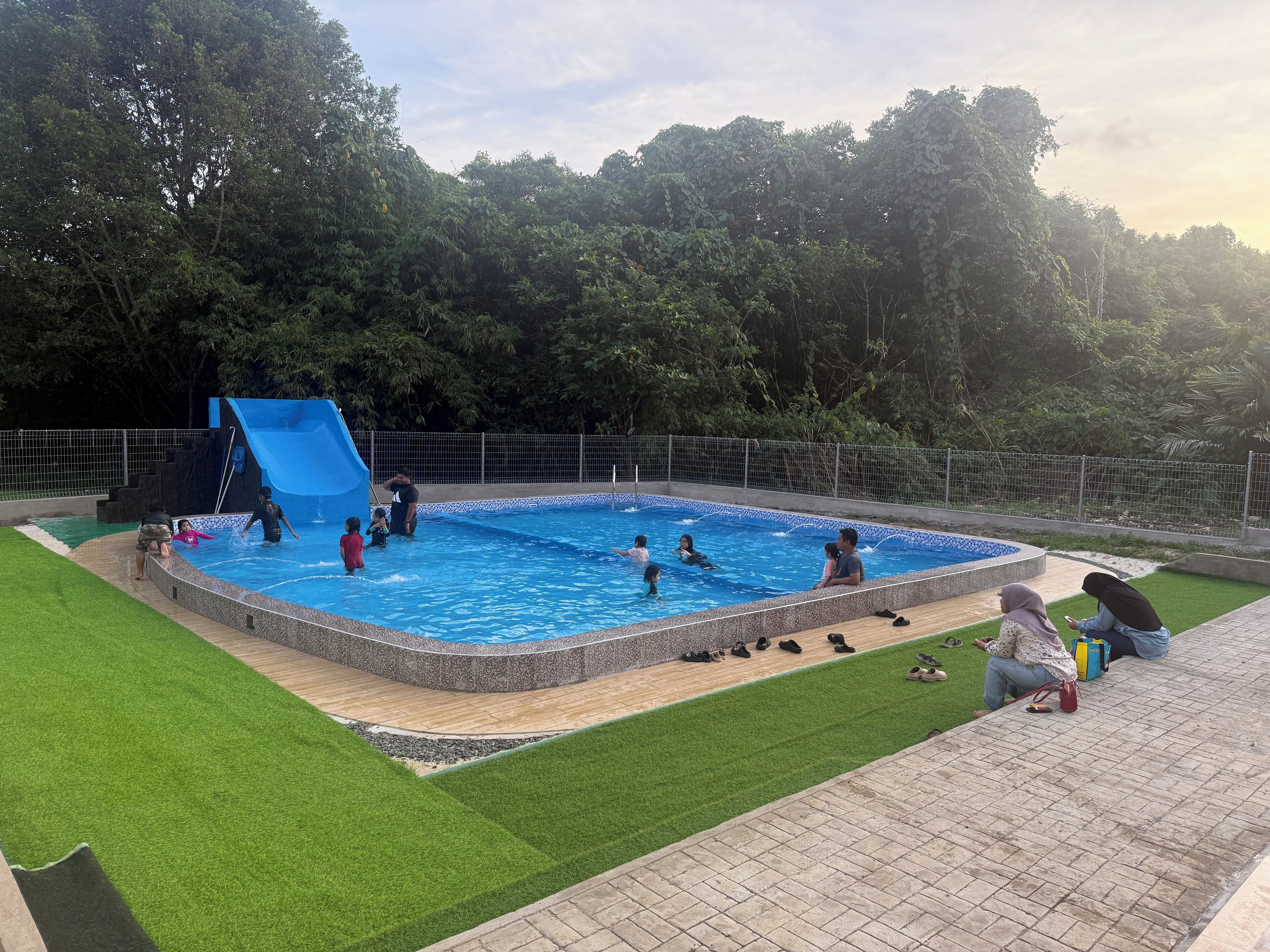 A swimming pool with a slide, surrounded by a fenced area and lush greenery.