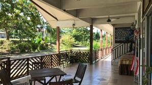 A covered outdoor seating area with wooden tables and chairs, a view of a garden with trees and a pathway, and a staircase leading to an upper level. - RTMS Guesthouse Semporna (Semporna)