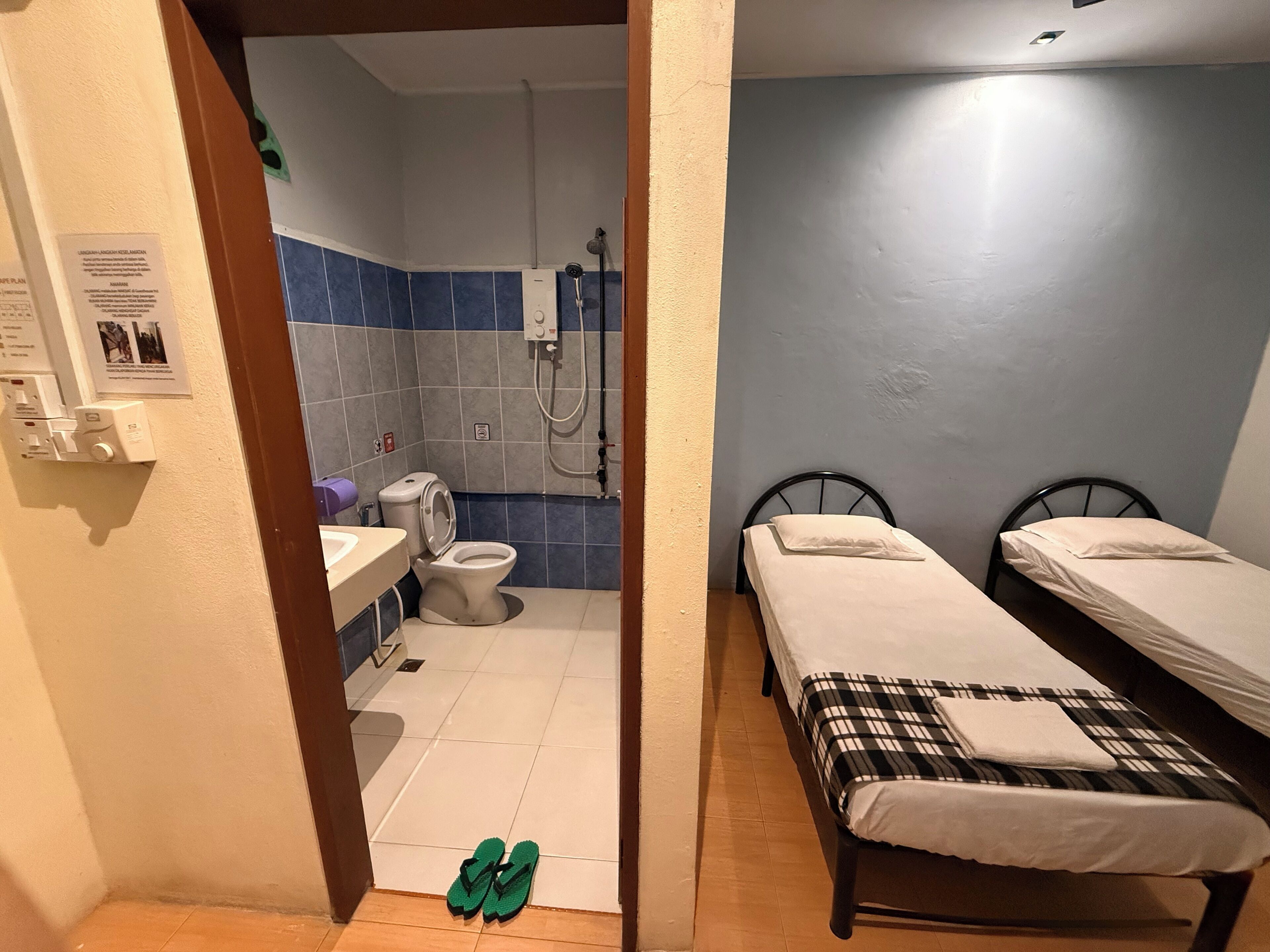 A small, compact room with a bathroom, two single beds, and a pair of green slippers on the floor.